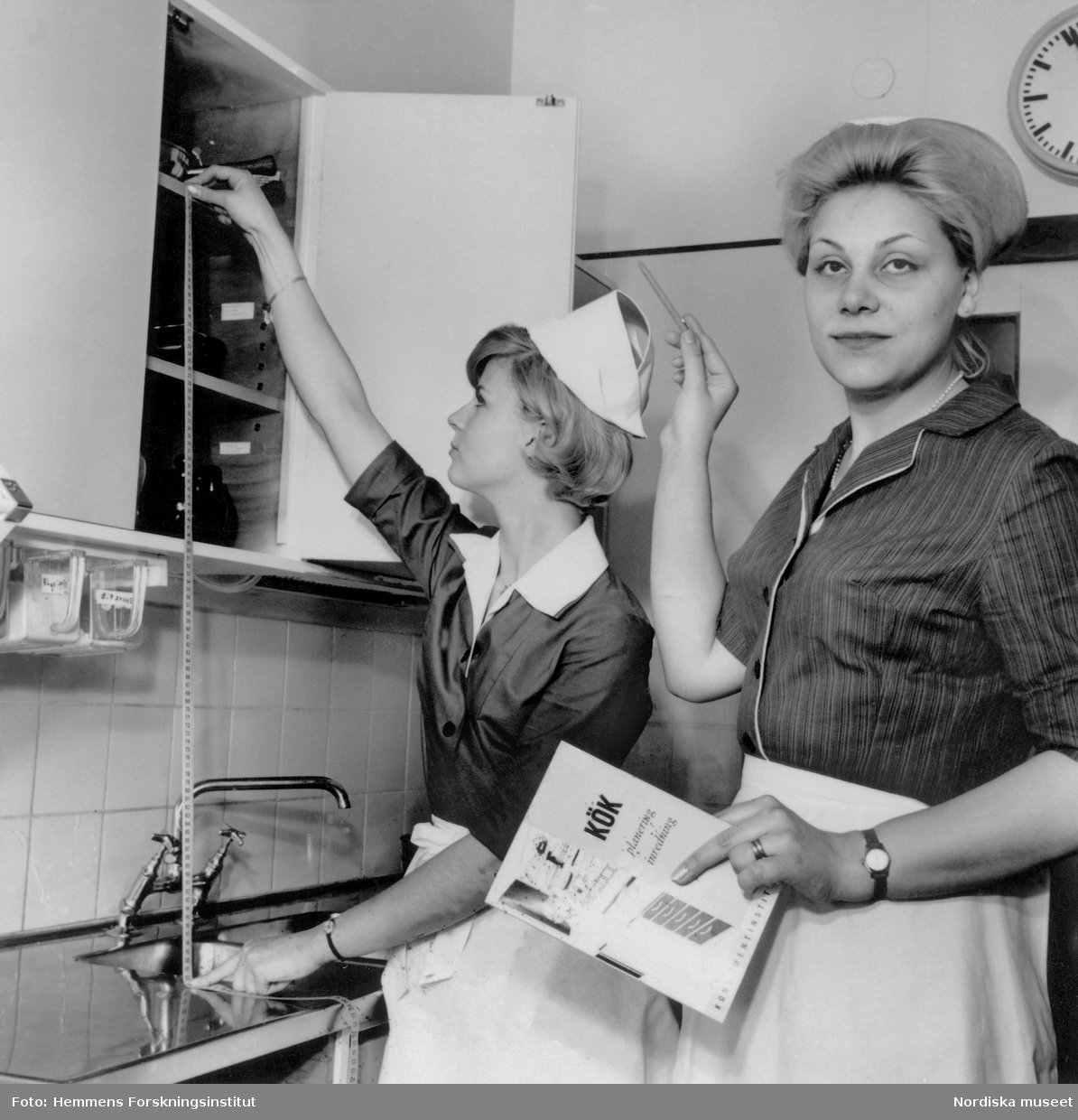 Två kvinnor mäter i ett kök. ”Kritiken mot 30-talets små kök var stark. Det blev inte som arkitekterna trott - att framtidens kök mest skulle användas för att värma färdiga konserver. På 40-talet försökte man ta reda på hur behoven verkligen såg ut. Hemmens forskningsinstitut som bildades 1944, koncentrerade sig främst på köket och utförde funktions- och utrymmesstudier i full skala. Med utgångspunkt i dessa studier fastställdes den första svenska köksstandarden 1950 som i stort sett är densamma som i dag.”