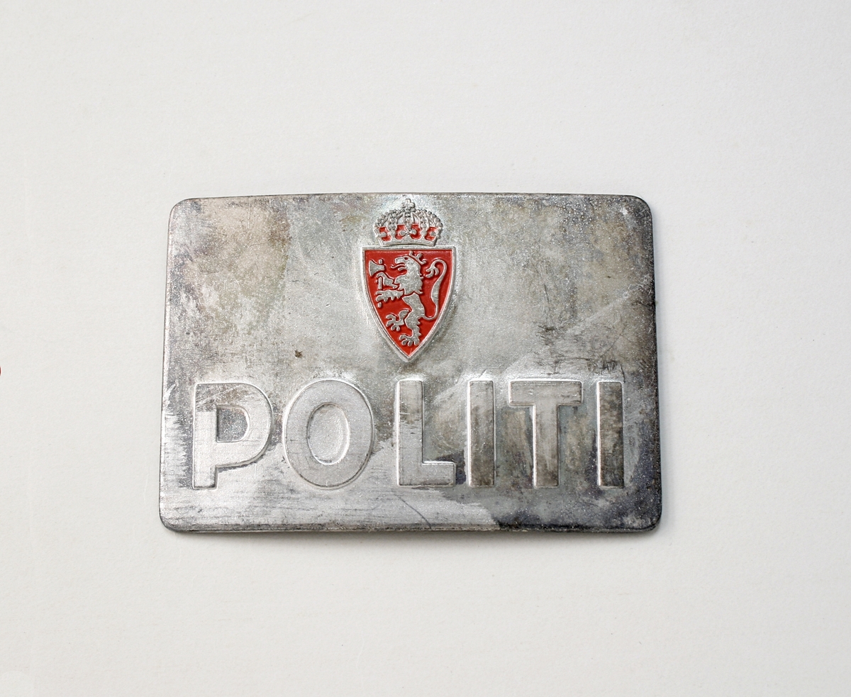 Rektangulært metallmerke med festenål på baksiden. Riksvåpen i rødt over ordet POLITI i relieff.
