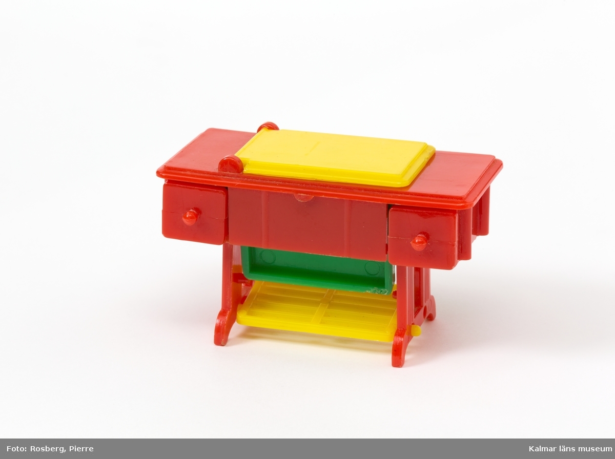 KLM 45437:7 Symaskin, av plast, dockskåpsmöbel. Bord i vilket en symaskin är nedsänkt. I det röda plastbordet finns en gul lucka som man lyfter upp och under den finns en symaskin som kan vikas upp. Symaskinen är i röd och grön plast. I bordet finns två lådor och en gul trampa/pedal.