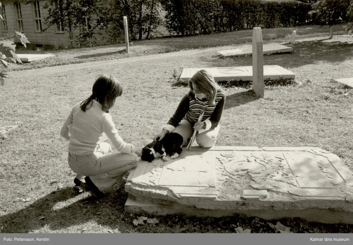 En katt som blir klappad på en gravhäll av ett par kvinnor i Gamla kyrkogården i Kalmar.