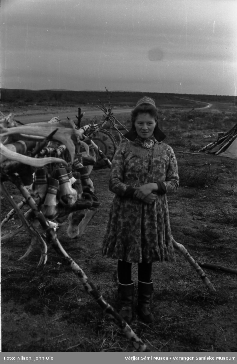 En ukjent kvinne i blomstrete sommerkofte selger duodji ved en hovedvei. Til høyre ses deler av en lavvo og til venstre et langt stativ av bjørk med reinhorn og skaller. Ukjent sted, Juli 1967.