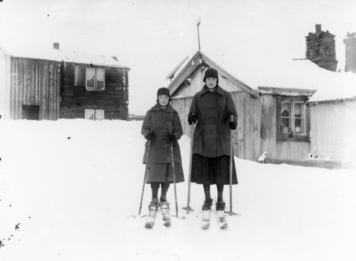 To jenter i skjørt, på ski foran to små hus, først på 1900-tallet