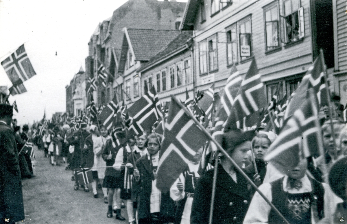 17. mai tog i Haraldsgt. Tilskuere på fortauet. Barnetog med norske flagg. Hull etter stifter i hjørnene.