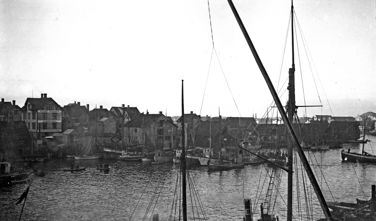 Havnbilde. Fiskebåter i havn. Mastene til et seilskip i forgrunnen. Bebyggelsen på Risøy og Bakerøy i bakgrunnen. Hverdagsfoto.