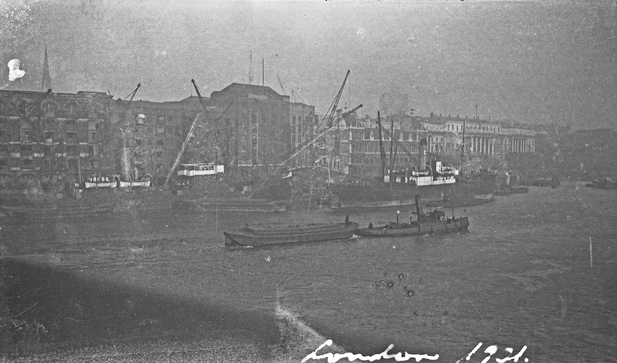 London havn. Flere skip ved kai. En taubåt med last på vei opp elven. Høye bygninger i bakgrunnen. Hverdagsfoto.