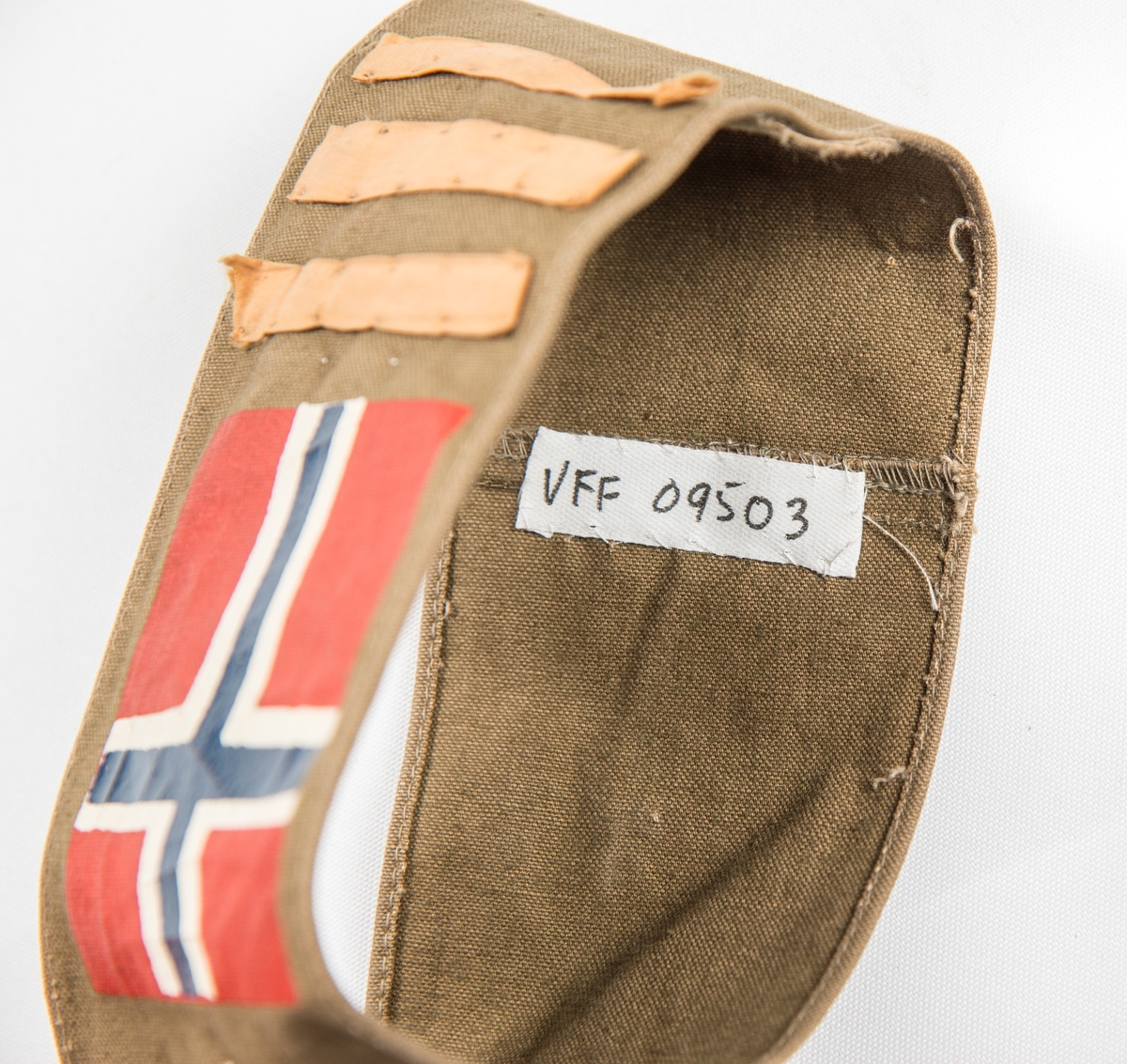Armbind (Kaptein) i brunt bomullstøy. Påtrykt det norske flagget og påsydd tre tynne gule striper. Var sett på ermet til VFF 09539 Uniformsjakke (med knappenål).