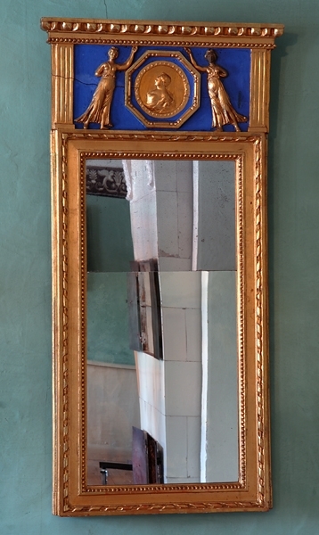 Hög spegel med 2- delat, rektangulärt glas. Förgylld, rikt profilerad träram med pärlstavsram närmast glaset och utanför denna en repstavsram. Spegeln har även ett blått överstycke med kannelerade pilastrar vid sidorna med ett krön av pärl- och äggstav. I det blå fältet dekor i förgylld relief bestående av ett medaljonporträtt i en åttakantig ram med en kvinna stående på vardera sida.