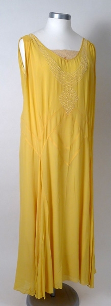 Lång sidenklänning av gul georgette, broderad med vita pärlor. Spets i urringningen både fram- och baktill på klänningen. Underklänning av gul crepé  de chine.