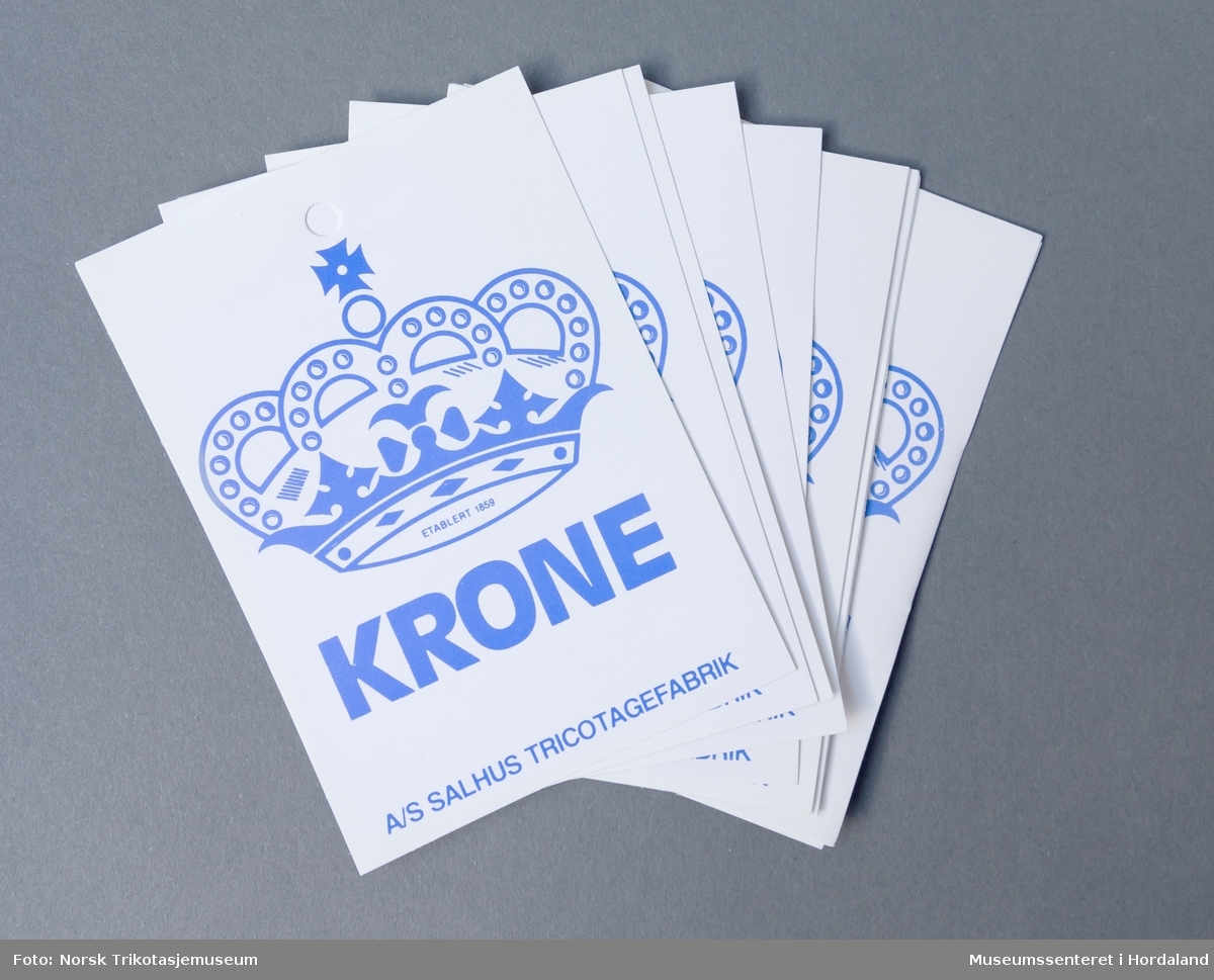 Merkelapp til plagg i papir, med poduktinformasjon og vaskeanvisningar, til plagg i merket Krone. Dette var det mest kjende merket til Salhus Tricotagefabrik.