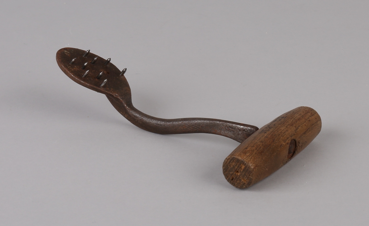 Sekkekrok/huk (lasteverktøy) i jern med ovalt endestykke med 9 spisse tenner i jern (1 stk. tann mangler). Med håndtak i tre.