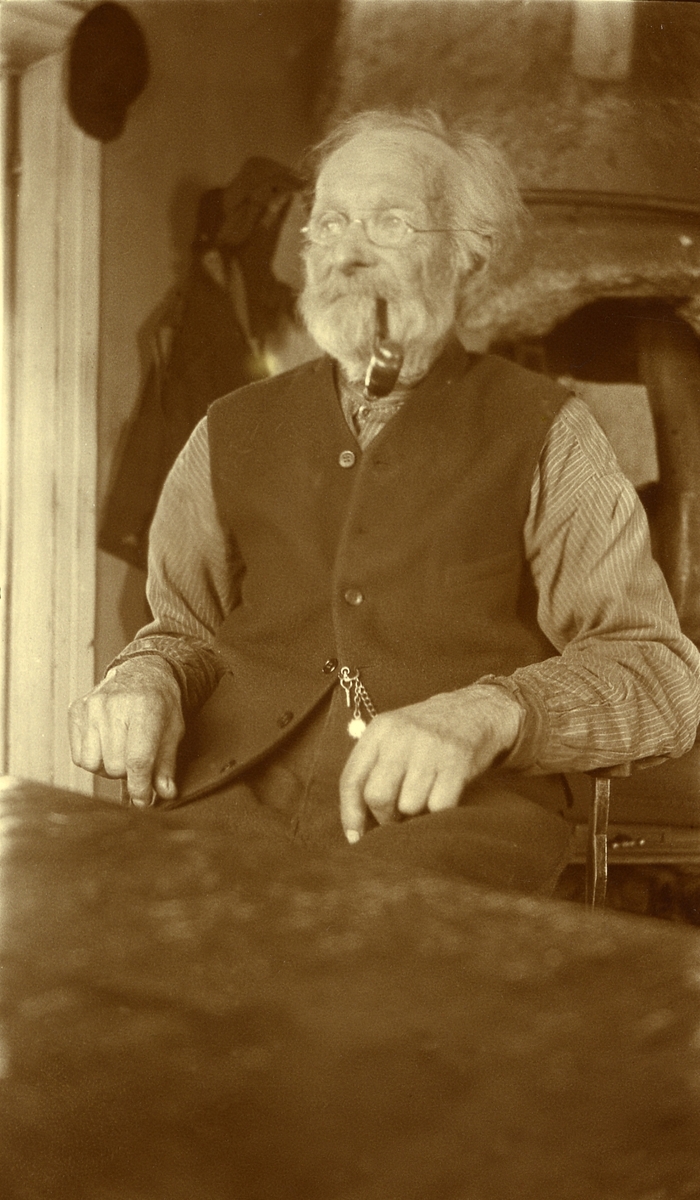 Mann sittende ved bord m/pipe i munnen i Langtjønnåsen, Sæter (?). Jon Olsen Sæter (Jonstø-Jon) (1845 - 1930)