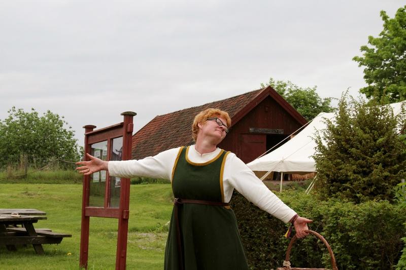 Rødhåret kvinne i middelalderdrakt, med kurv i hånda, slår ut med armene for å ønske velkommen til Urtehagen (Foto/Photo)