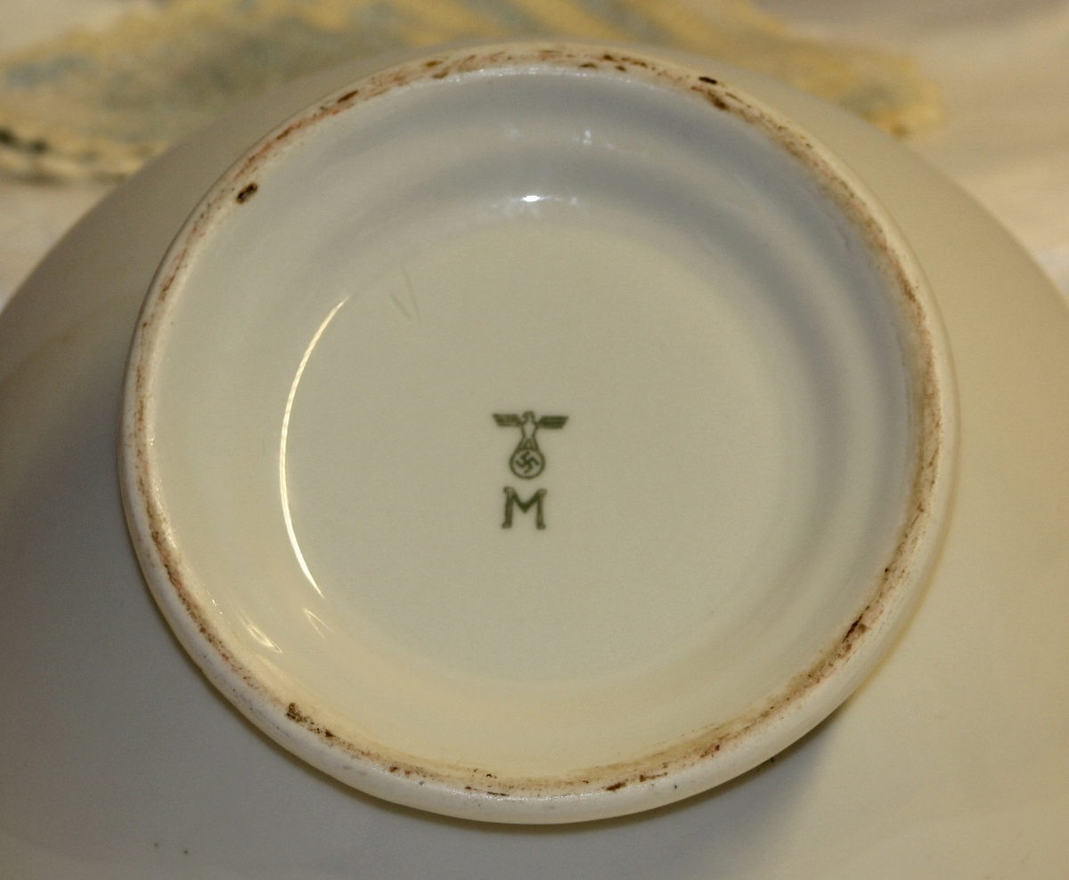 Rund suppeterrin av hvitt porselen på rund fot. Den har to løvehoder som håndtak. Under bunnen er et nazisymbol med en ørn som bærer et hakekors over bokstaven "M".