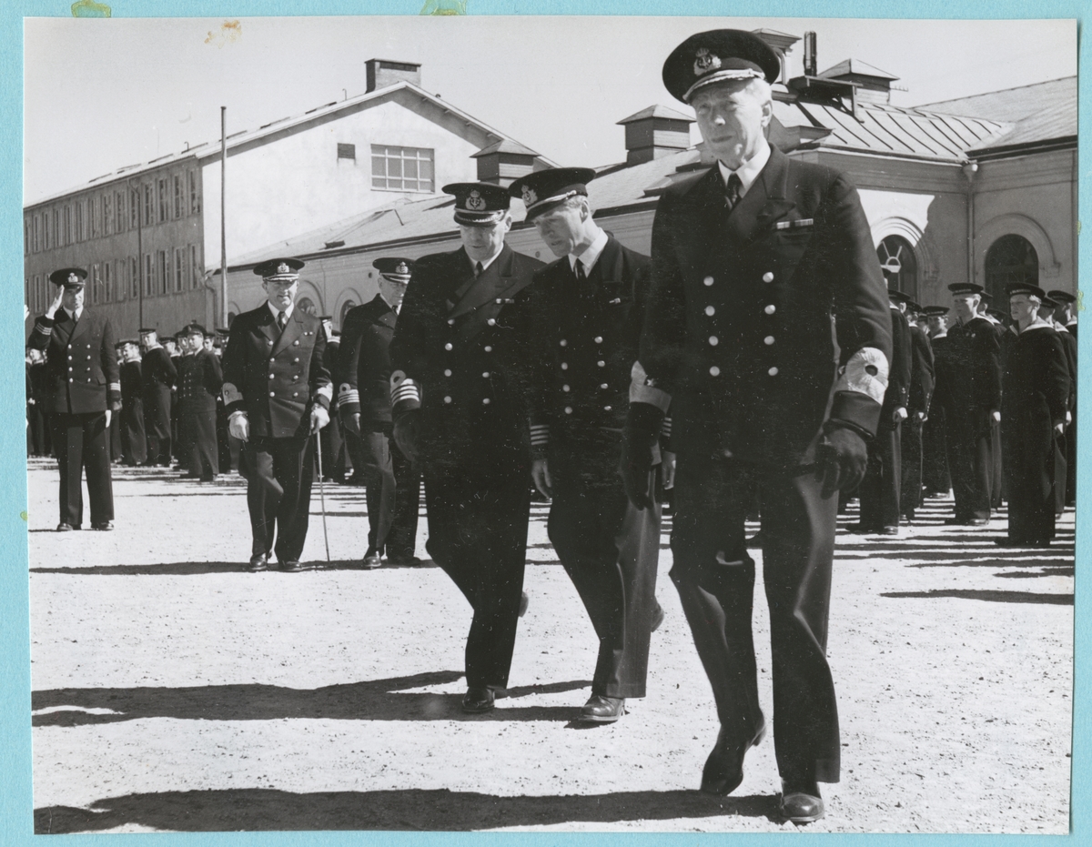 Viceamiral Strömbäck och konteramiral Samuelson går tillsammans med en grupp höga militärer på bataljon Sparres kaserngård. Bakom dem står flottister uppställda. Samtliga är klädda i uniform. I bakgrunden syns Sparres exercishus.