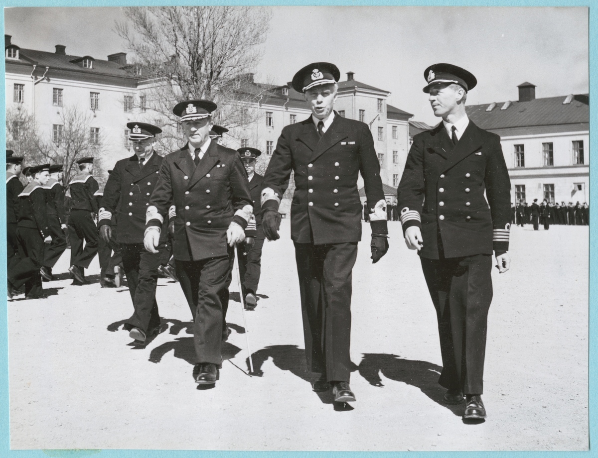 Viceamiral Strömbäck och konteramiral Samuelson går tillsammans med en grupp höga militärer över bataljon Sparres kaserngård. Bakom dem marscherar en grupp med flottister förbi. Samtliga är klädda i uniform. I bakgrunden syns kasern Sparre.
