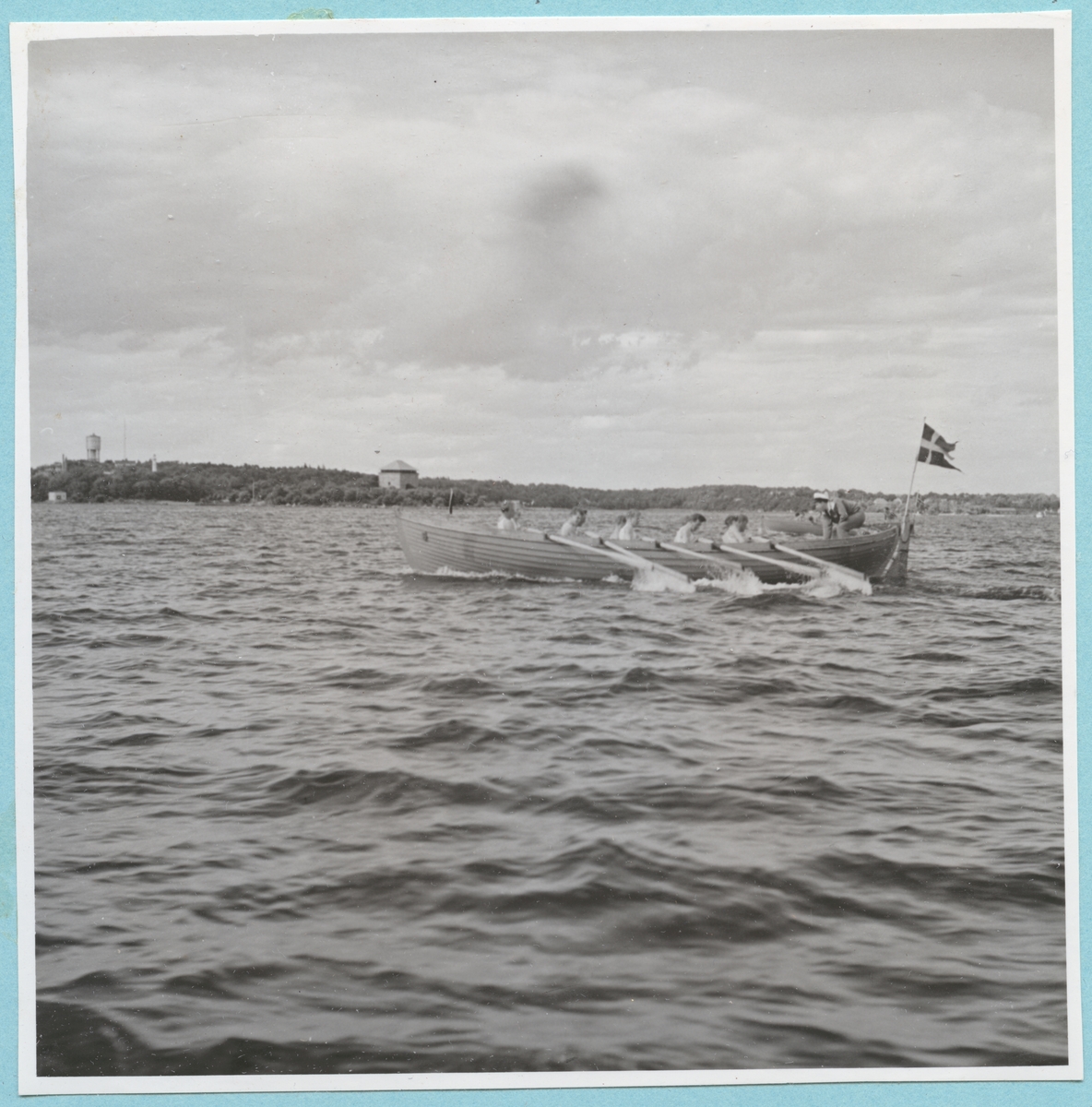 Roddtävling mellan Danska, Norska och Svenska kadettlagen. Från Danska och Norska örlogsbesöken den 29/6 - 4/7 1951. Flera män sitter i en roddbåt och ror. I aktern av båten sitter en flagga. I bakgrunden syns Mjölnareholmen med kruthuset och Wämö med vattentornet.