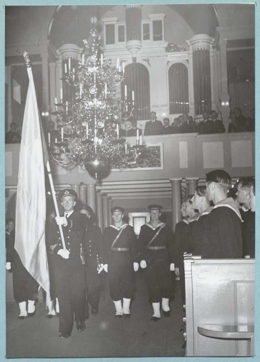 Bilder från Krigsmans erindran den 13/10 1951. Uniformsklädda flottister går genom Amiralitetskyrkans mittgång, över vilken det hänger en ljuskrona i mässing. Längst fram går en löjtnant som håller i en flagga. I bänkraderna syns ytterligare flottister stå upp. I bakgrunden syns kyrkans orgelläktare som bärs upp av doriska kolonner. På läktaren, framför orgelfasaden, står flera män.