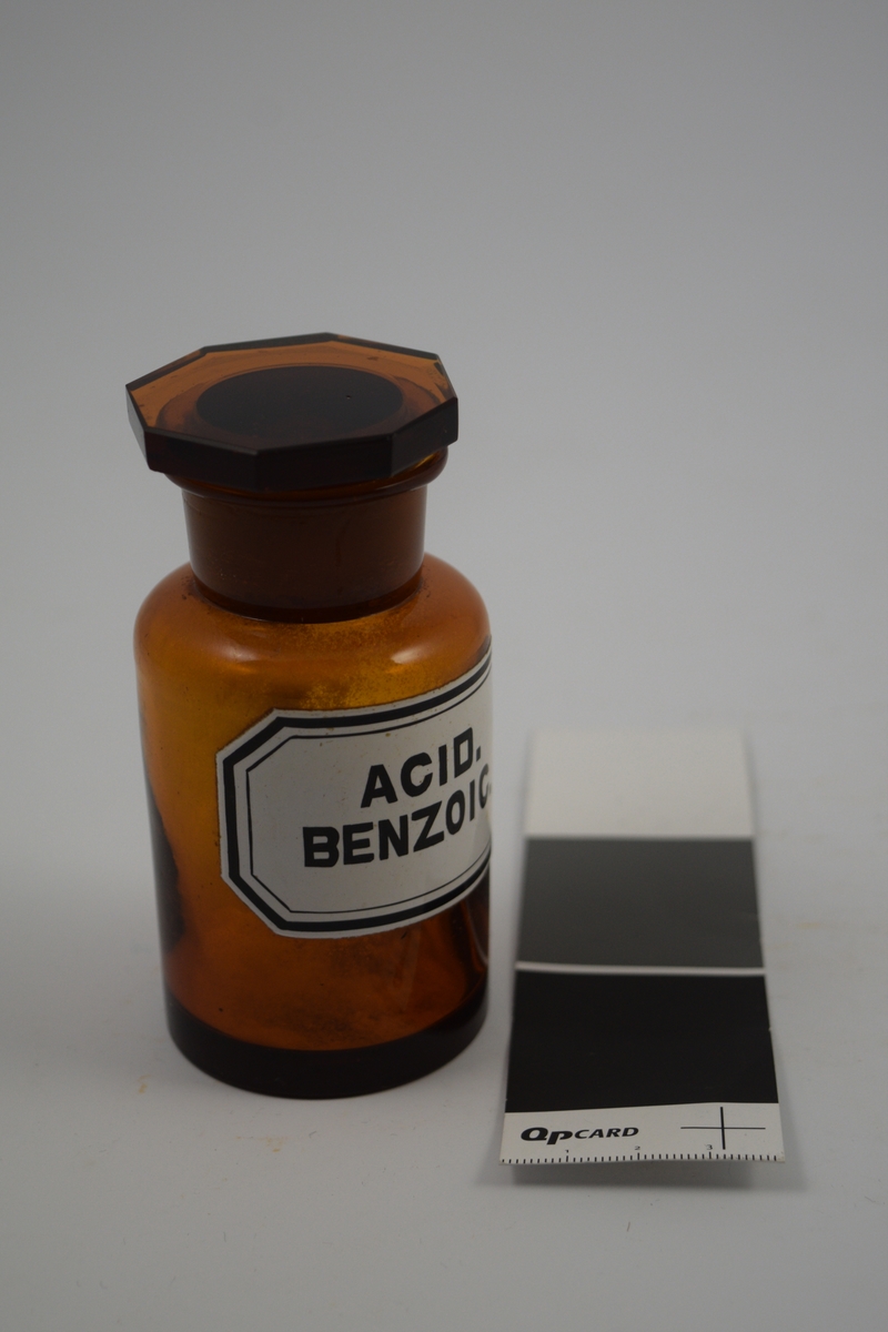 Brun glasskrukke med glasspropp. Hvit etikett med sort skrift. Acid Benzoic ble brukt som konserveringsmiddel til salver og øyedråper med mer. Også brukt til syltetøy.