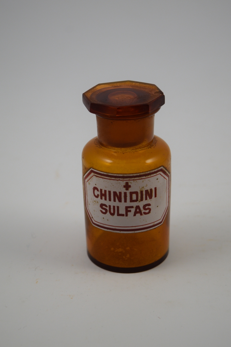 Brun glasskrukke med glasspropp. Etikett hvit, med rød skrift. Ett kors, betyr at innholdet er giftig. Chinidini Sulfas ble brukt i hjertemedisin.