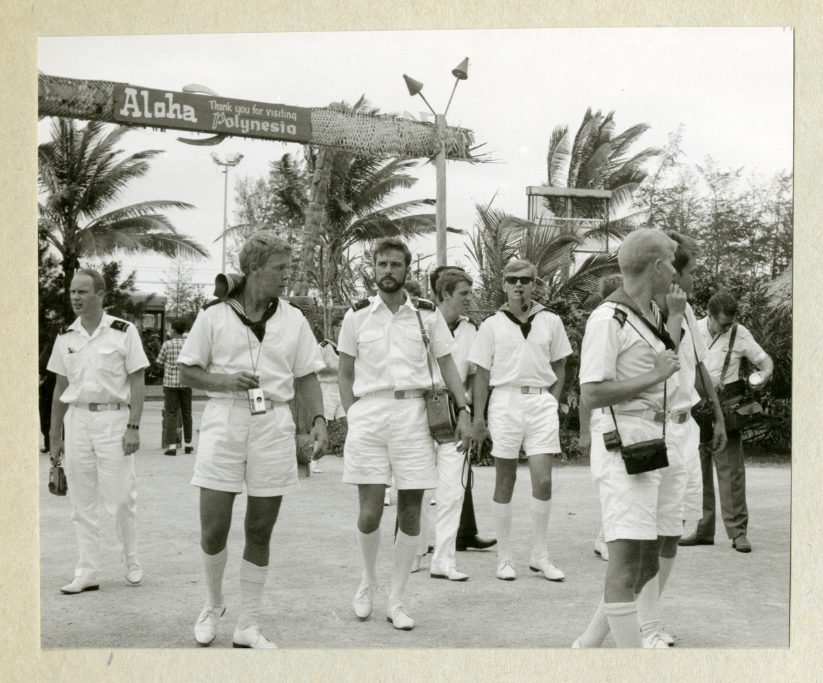Bilden föreställer en grupp besättningsmän i vita sommaruniformer på utflykt till Waikiki beach. I bakgrunden syns palmer och en "Aloha"-skylt. Bilden är tagen under minfartyget Älvsnabbens långresa 1966-1967.