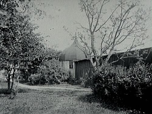 Trädgård med sex- eller åttakantligt (lust)hus med liten spira som står invid en långsträckt byggnad.