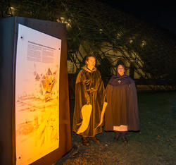 To kvinner i middelalderklær og kapper står foran en svakt opplyst middelalderruin og synger.
