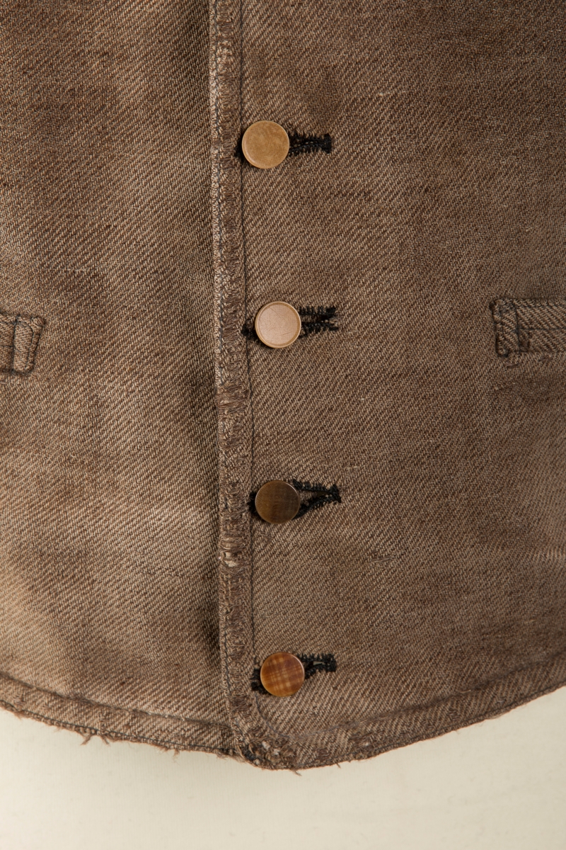 Form: Forstykker av ull, rygg av bomull. 7 knapper av 5 ulike slag, to små lommer uten klaff. Spennetamper på ryggen.