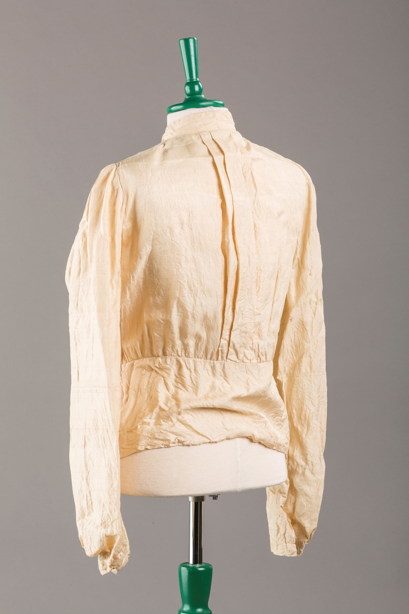 Bluse med sløyfe i halsen og kappe nederst, 9 trykknapper, leggsying på ryggen.
