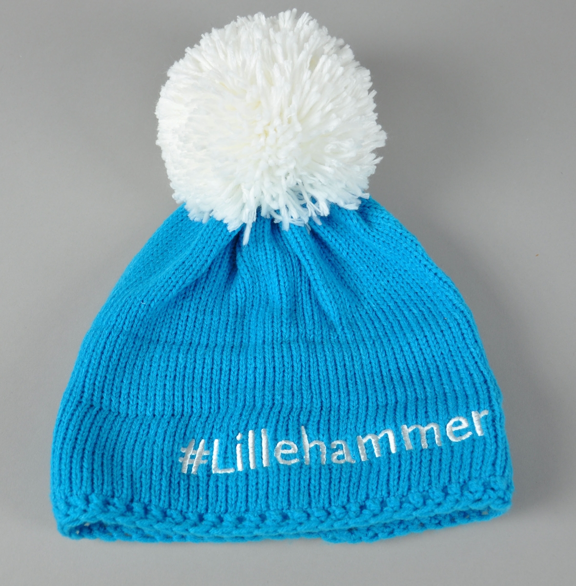 Lyseblå strikket lue med hvit dusk. Påsydd logo for Lillehammer Youth Olympic Games 2016.