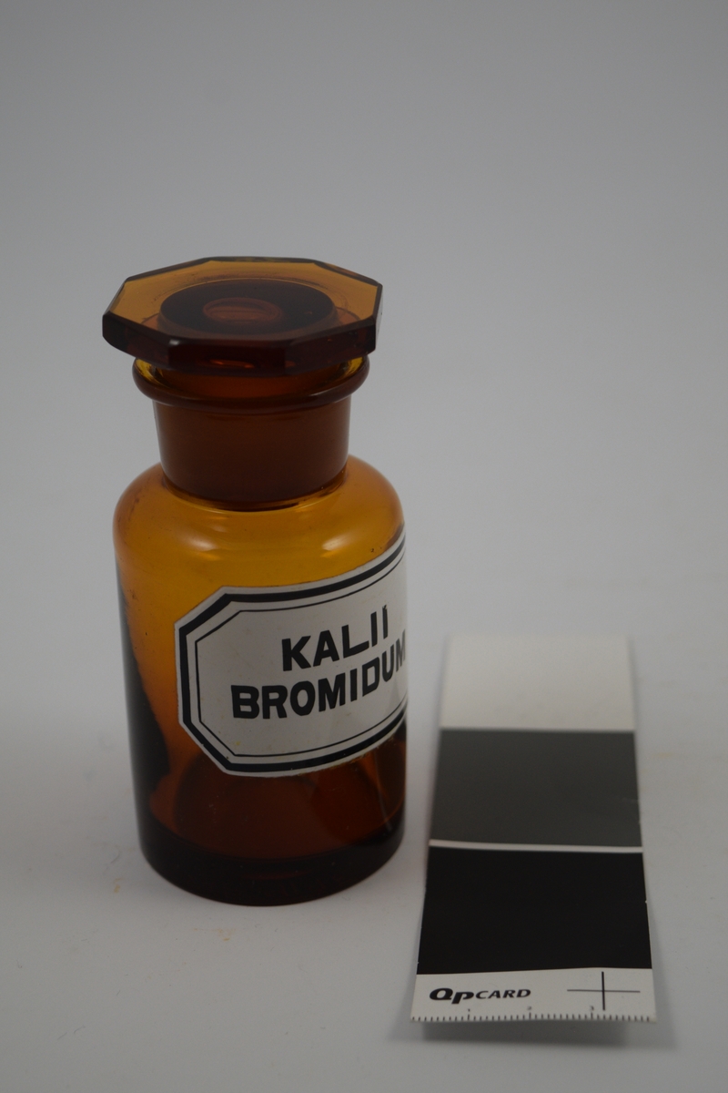 Brun glasskrukke med åttekantet glasspropp. Hvit etikett med sort skrift. Krukken ble brukt til oppbevaring av Kalii bromidum, som ble brukt som sovemiddel.