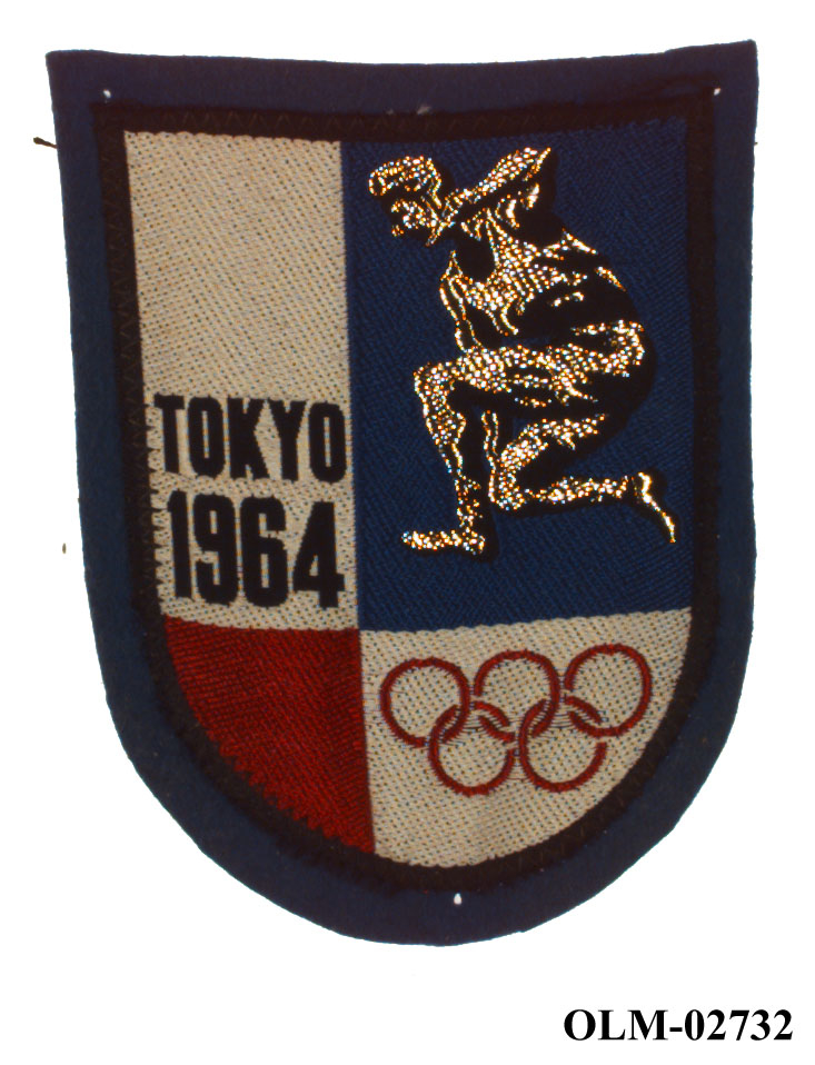 Skjoldformet tekstilmerke fra sommer-OL i Tokyo 1964. Merket har mørkeblå kant og er delt i fire deler, en for sted, en med friidrettsutøver, en med de olympiske ringer og en del er rød.
