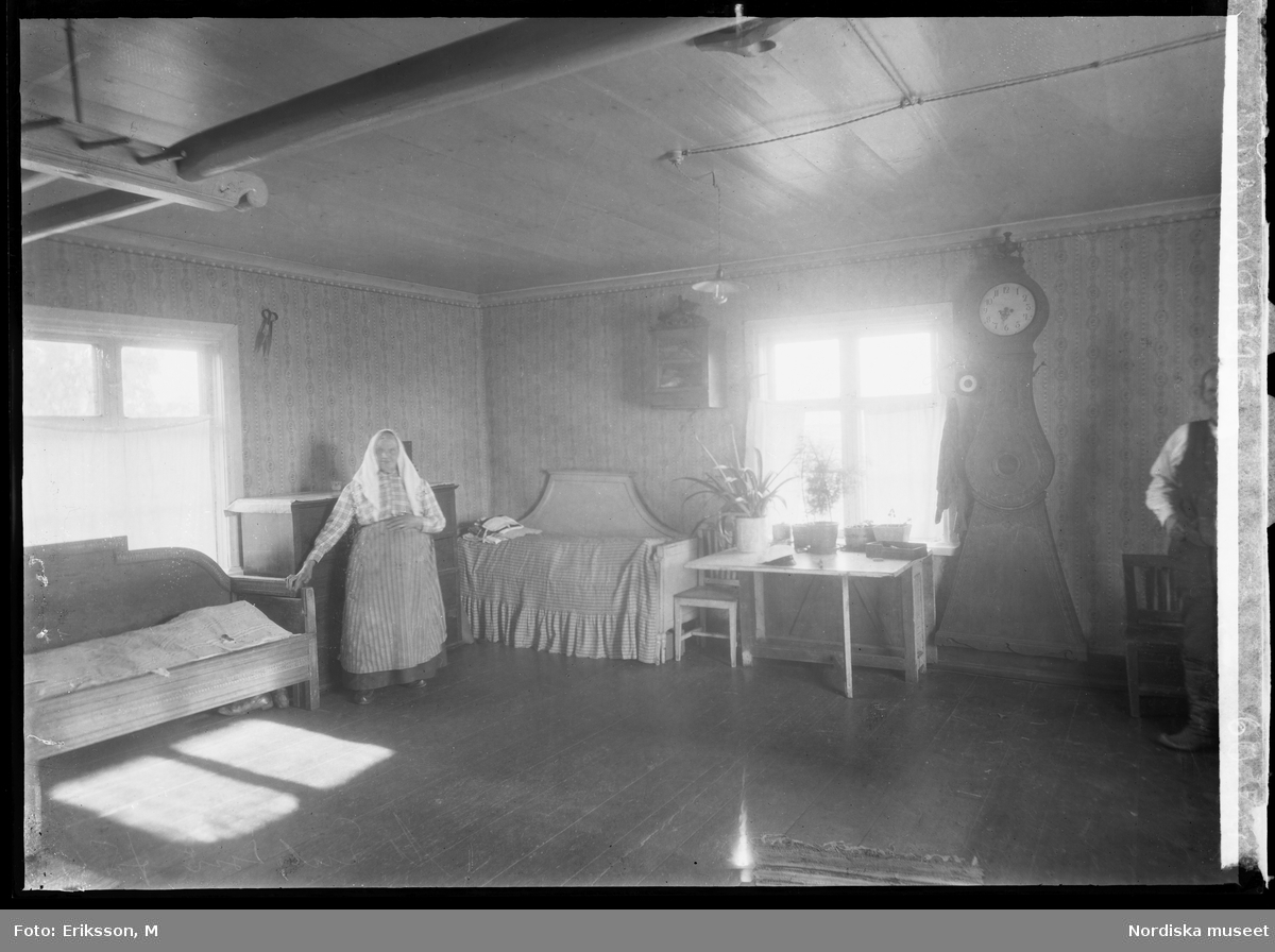 Interiör från pörtet. En kvinna står och håller sig i en av de två sofforna som står längs väggarna. Vid fönstret ett skrivbord och ett golvur.