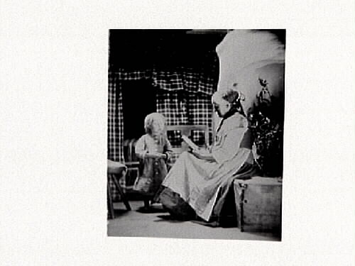 Arrangerad bild. Kvinna iklädd folkdräkt läser för ett stående litet barn.