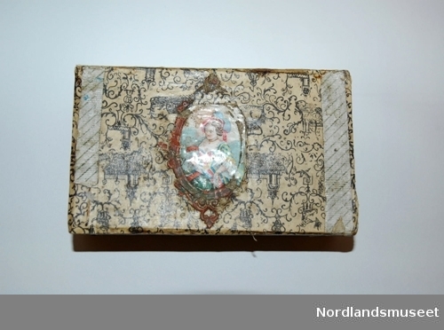 Rektangulært smykkeskrin laget av tre. Utsiden er dekket av ornamentert papir. Kan være gavepapir. På lokket er det også limt på et glansbilde med bilde av en dame.