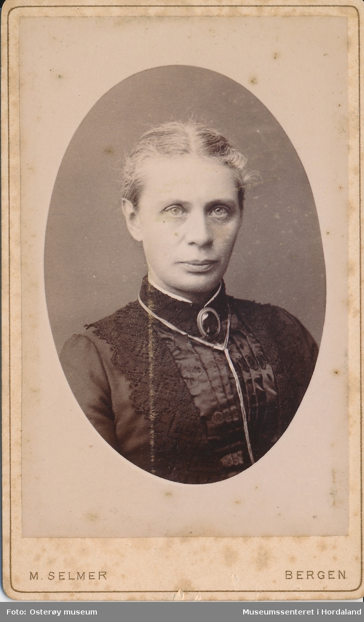 portrettfotografi av kvinne med midtskill og håret samla i nakken, kledd i mørk kjole med blonder og silke i brystet, brosje og kjede i halsen