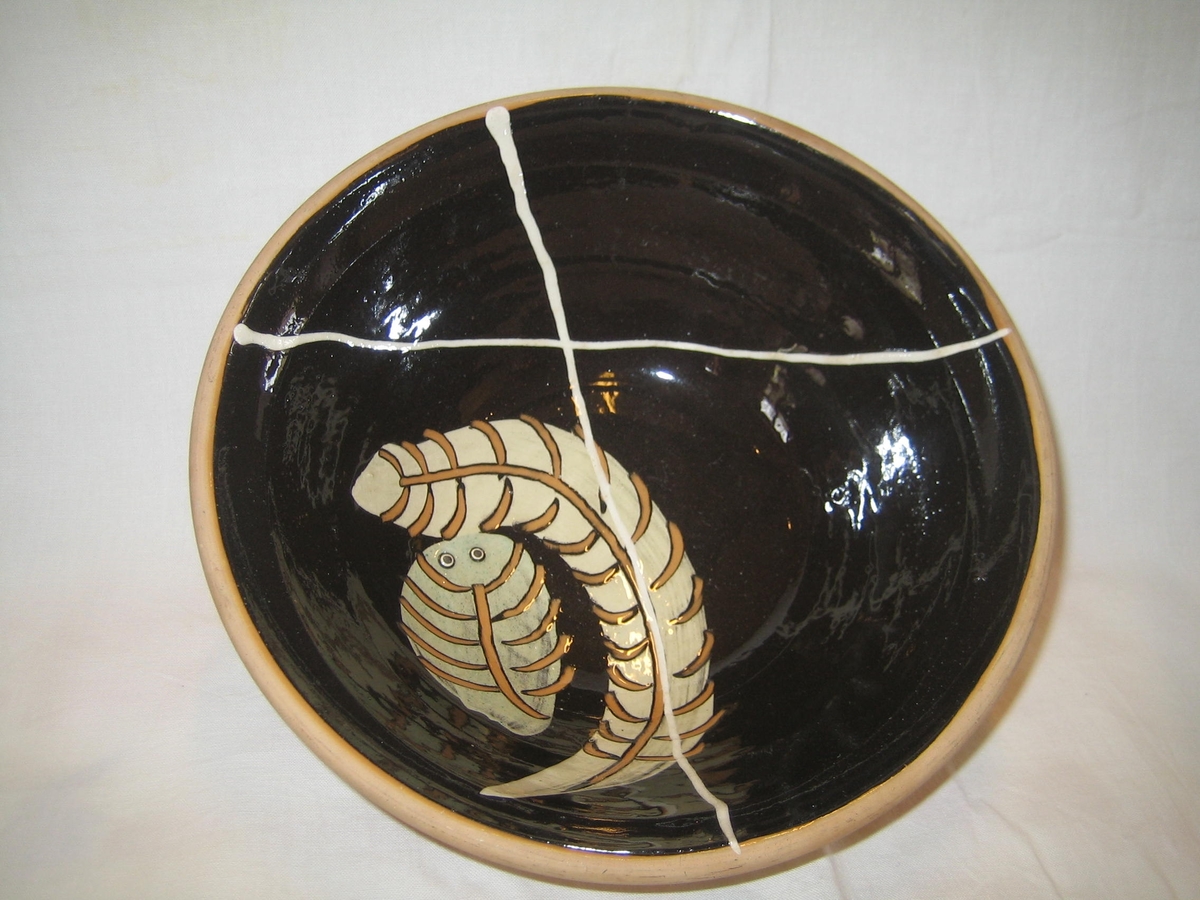 Rund skål i naturfärgad keramik på utsidan och svart glasyr på insidan, dekorerad med fjädrar/blad i orange och vitt.
