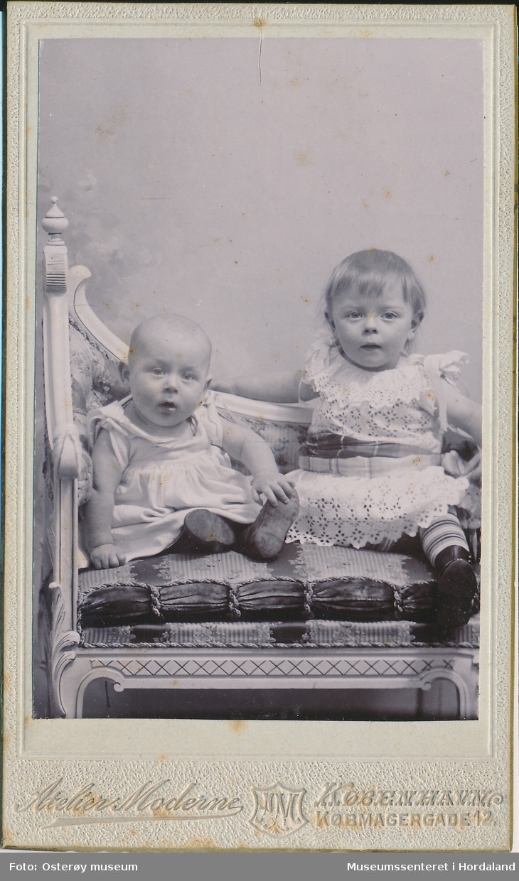 atelierfotografi av to små born, gut og jente som sit i ein stol kledd i ljose kjoler