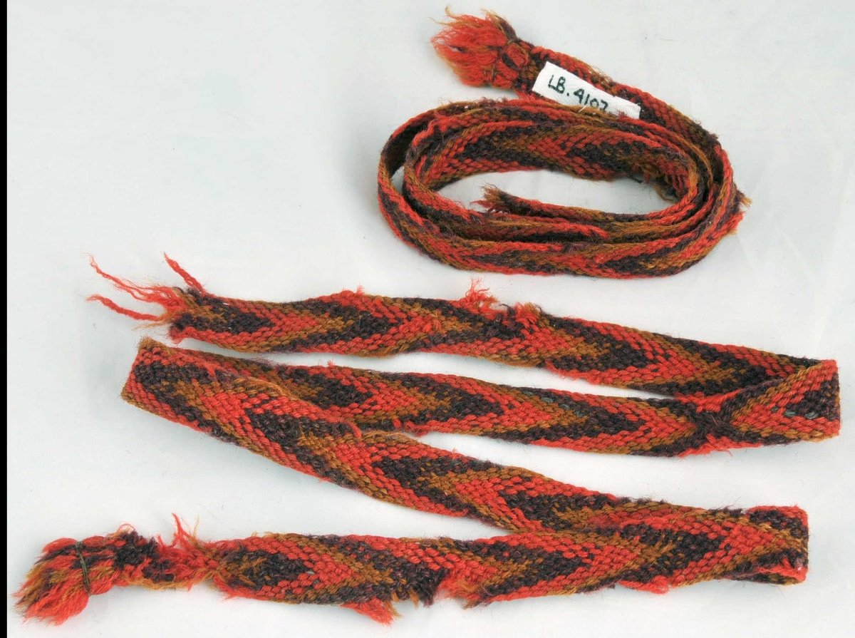 Slengde sokkeband laga av 8 trådar raudt, 8 trådar fiolett og 6 trådar okegul. Små frynser i begge endar.