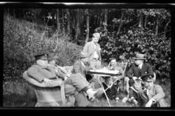 Gruppe av unge og gamle menn samlet rundt et bord i hage. Fr