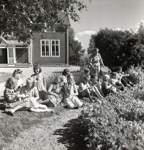 Veckebobarnen, Unga Odlare i Ovanåker 1950. En grupp barn sitter ute i gräset. Solen skiner. En kvinna serverar dem dryck med skopa ur en hink.