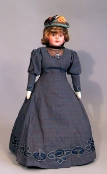 Docka med celluloidhuvud sydd omkring 1970 med kläder i 1890-talets stil. Blå-brun klänning och sex underkjolar, mamelucker samt kängor i ljust skinn.
