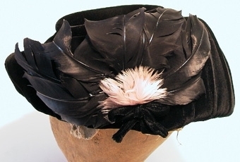 Damhatt sydd av svart sammet.Sydd i s.k. barettmodell utan skärm.Tyget lagt i mjuka veck. Prydd i nacken med en rosett av sammet samt svarta och rosa fjädrar.