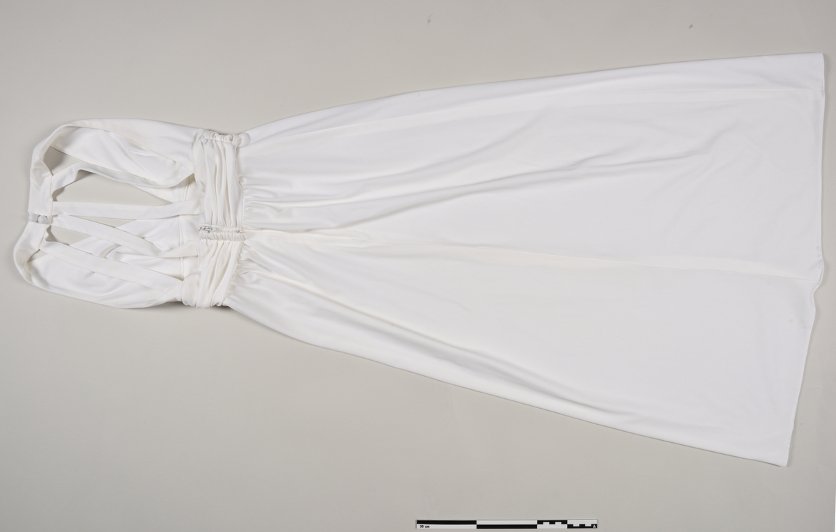 Kremhvit kjole sydd av polyesterstoff. Det er en glidelåsåpning bak og 5 pynteknapper foran. Kjolen har en V-utringning og ingen ermer.