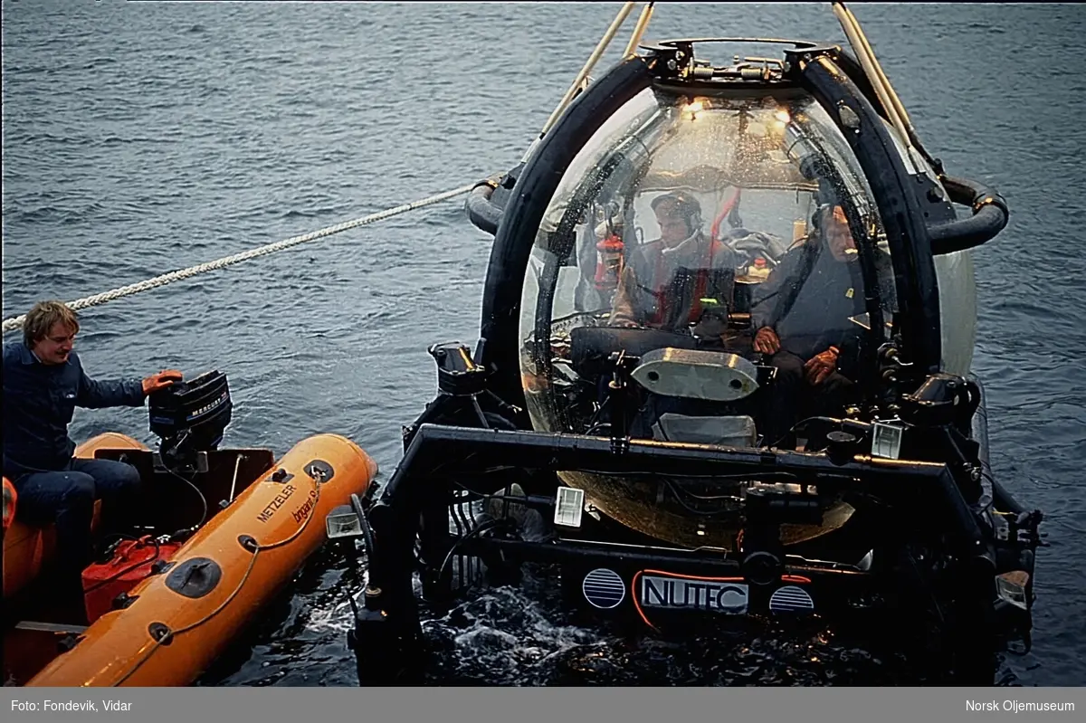 To menn i en liten gummibåt kjører inntil NUTEC's miniubåt Check Mate, som holdes rett over vannoverflaten i Gravdalsbukten. To menn sitter ombord i ubåten. NUI's dykkersenter ligger i bakgrunnen.