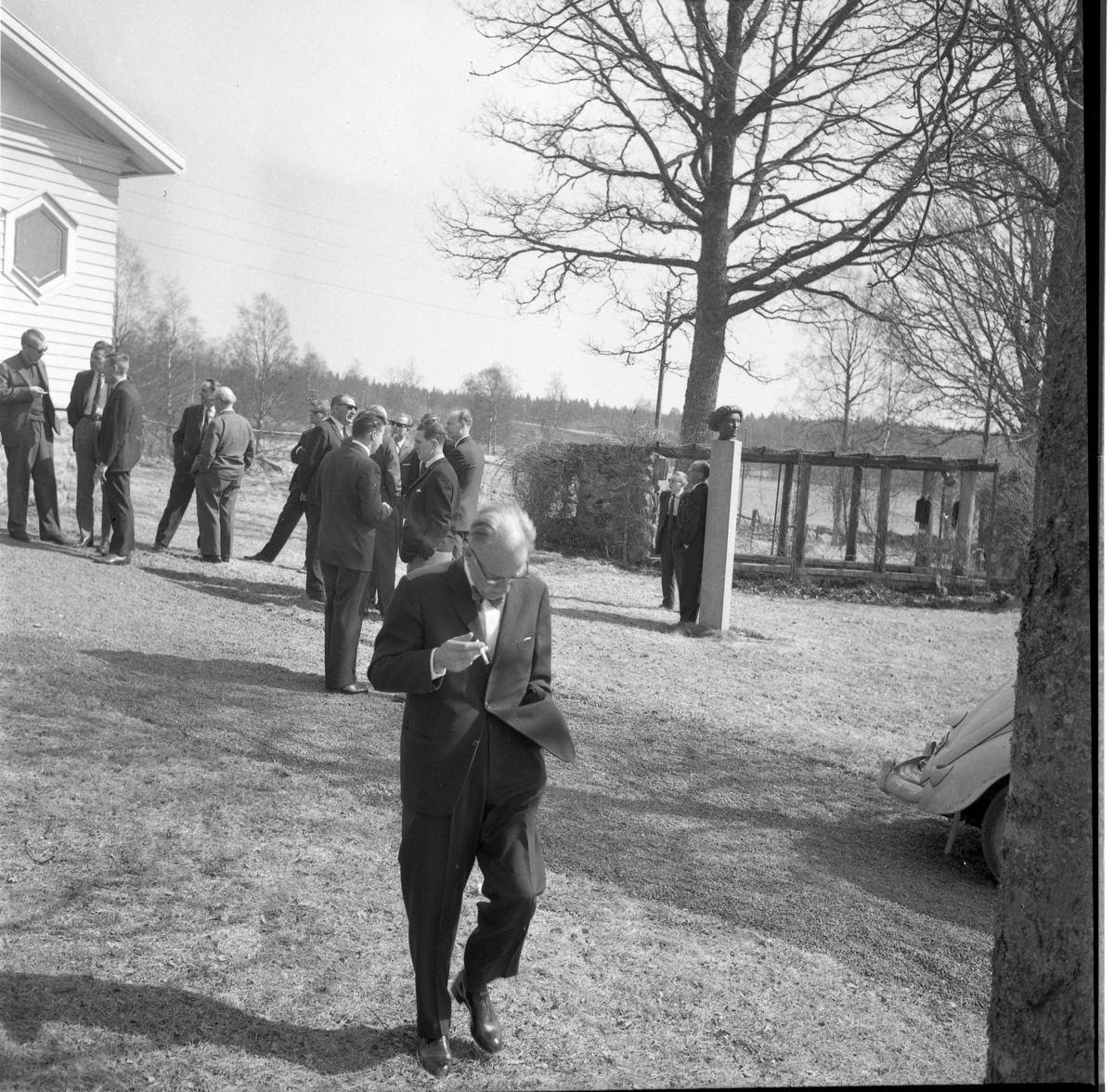 H Hall-konferens i Lövhult 1964. Okända kostymklädda män utanför en byggnad samlade i olika små grupper. En cigarettrökande med går med nedböjt huvud mot fotografen.