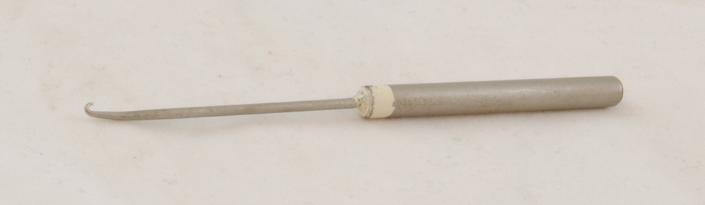 Spetskrok bestående av en metallpinne med en krok fastlött i ett handtag av metall. Verktyget är markerat med ett vitt streck runt handtaget vilket visar att det tillhör teleavdelningen på SJ-skolan, Ängelholm.