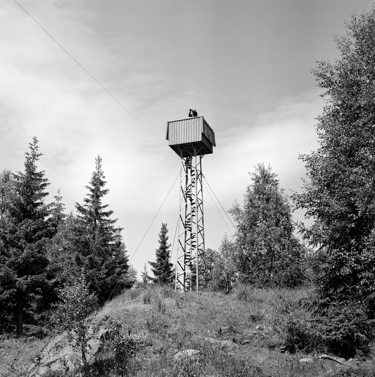Utsiktstårnet ved brannvaktstasjonen på Flotsberget i Elverum.  Fotografiet viser et smål jerntårn med en kasseformet utsdiktsplattform på toppen, med oppgang via ei innvendig vindelstrapp.  Den forholdsvis smale, høye konstruksjonen ble holdt i posisjon av barduner, jernvaiere som var forankret i fjellet omkring.  Da dette fotografiet ble tatt sto en mann med kikkert oppe i tårnet og speidet ut over det omkringliggende skoglandskapet.  Dette var antakelg skogbrannvakt Nils Granli (1903-1975).  Fløtsberget ligger om lag 560 meter over havet, øst for grenda Melåsberget i søndre del av Elverum kommune.  Den første skogbrannvaktstasjonen her ble etablert av lokale skogeiere i 1904.  Forsikringsselskapet «Skogbrand» trådte støttende til da anlegget ble modernisert i 1933.  Elverum kommune var eier da vaktholdet på Flotsberget ble avviklet etter 1973-sesongen.  Kommuneingeniøren foreslo da at peileapparatet, kikkertene og kartene som hadde tilhørt anlegget skulle overdras til Norsk Skogbruksmuseum.  Sjølve hytta er seinere gjort tilgjengelig for allmennheten som kvilested for folk bruker omkringliggende terreng i rekreasjonssammenheng. Branntårn.