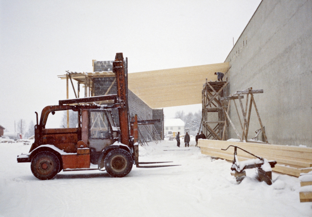 Fra sagbrukstomta ved Våler skurlag på Braskereidfoss i Solør.  Dette er et vinteropptak med snø på marka.  I forgrunnen sto det en truck.  Bak ser vi bygningsarbeidere i sving med å legge et slakt pulttak over et tilbygg til en murt industrihall.  Dette antas å være høvleriet, som ble bygd i 1967-68.