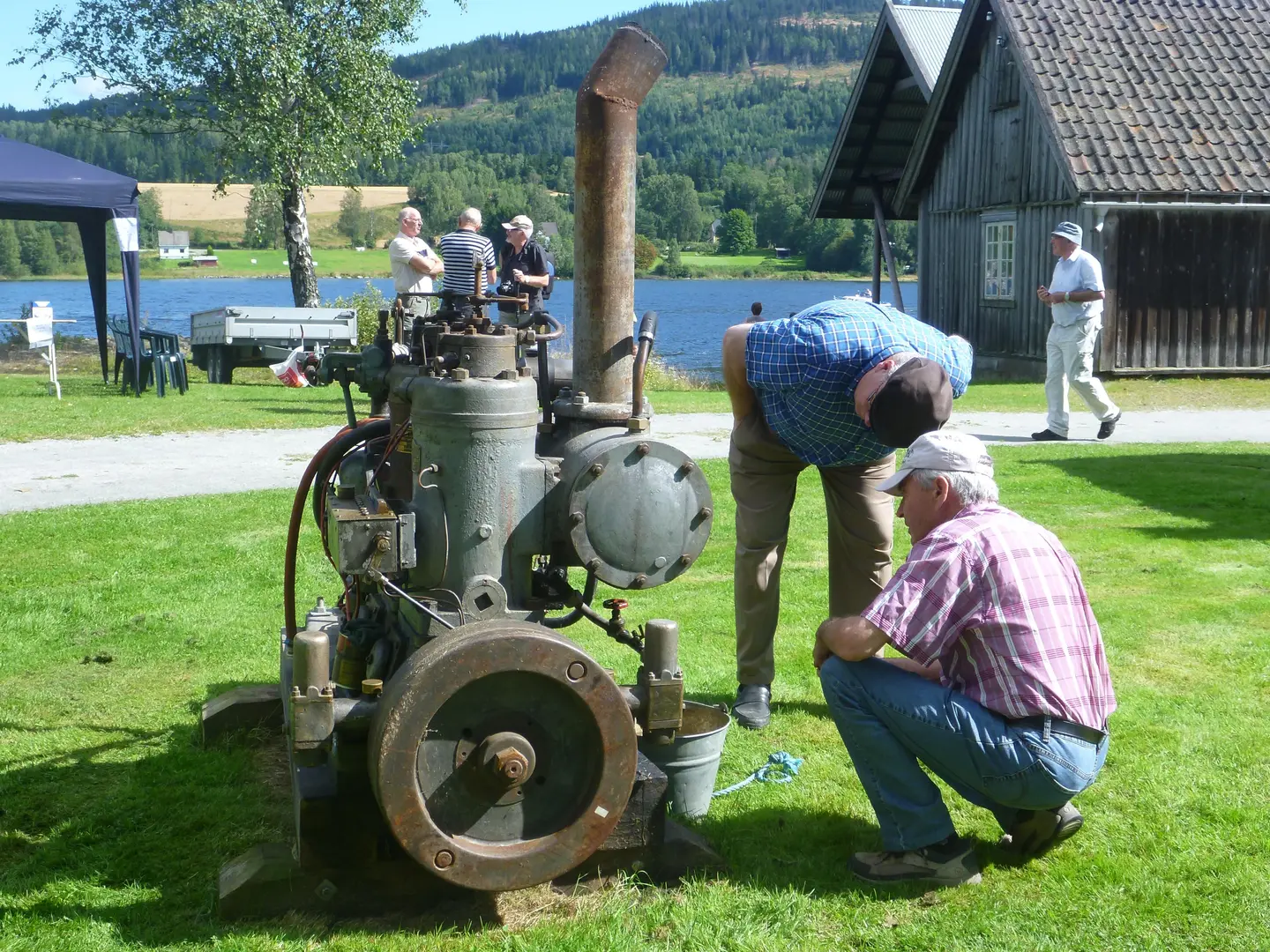 To menn ser på en gammel dieselmotor som står utendørs. I bakgrunnen Mjøsa, et gammelt trehus og grønn plen.