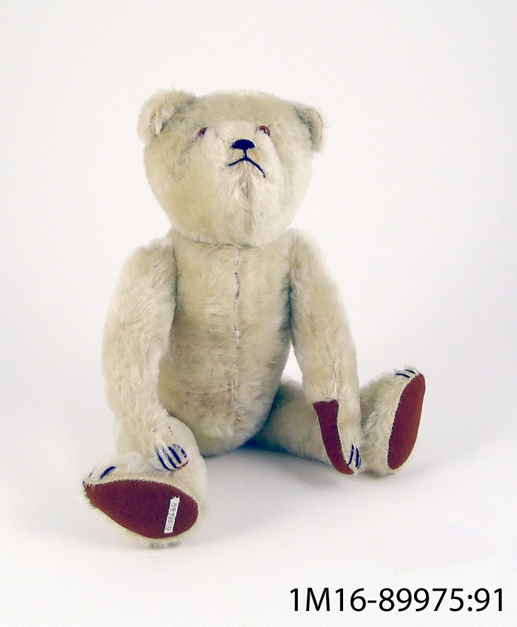 Teddybjörn av vit plysch med trampdynor av brun filt, klor av svart tråd, bruna ögon av glas samt mun och näsa i svart, defekt.